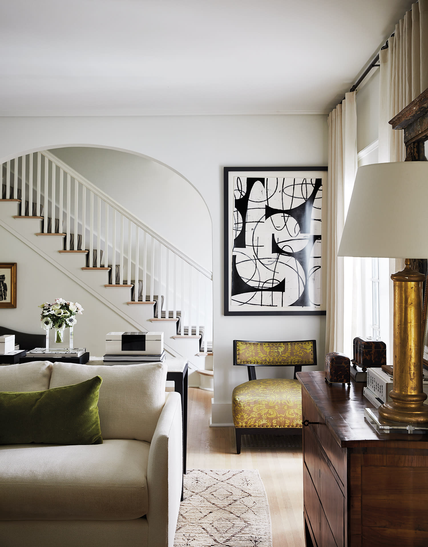Living room with arched stair opening, neutral sofa, abstract art, and vintage wood furniture.