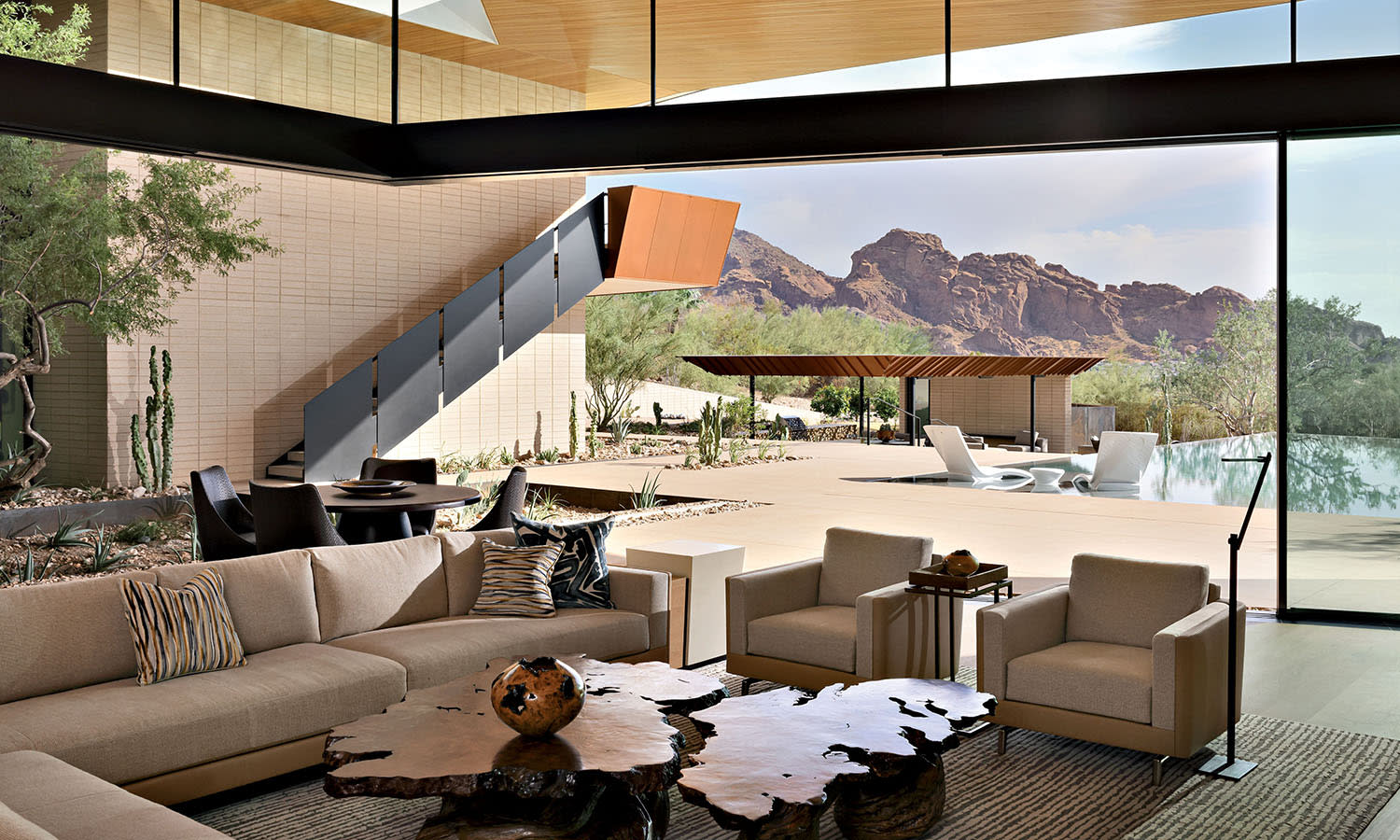 Modern living room with large glass walls showcasing a desert landscape. Features beige sofas, a wood coffee table, and a sleek staircase. Bright and airy.