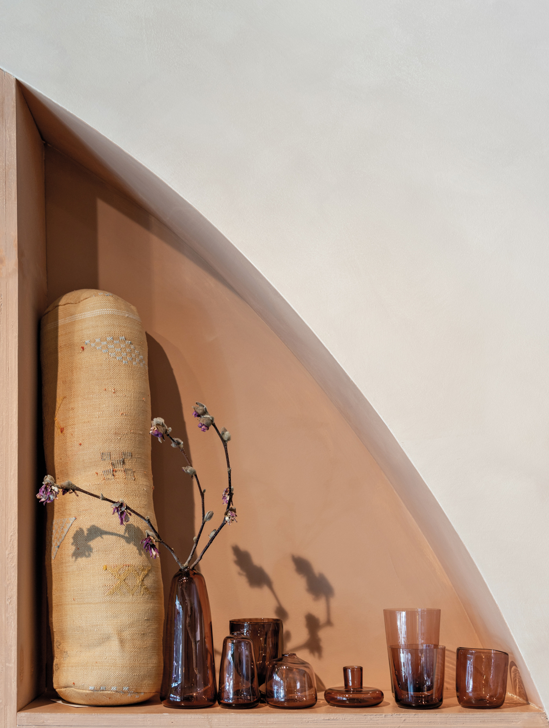 Arched shelf with embroidered bolster pillow and dark purple glass vessels at Origin Story