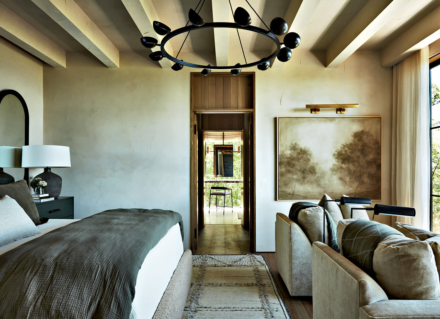 A bed made with white linens and a darker quilt sits with two chairs in front of it, all facing a window.