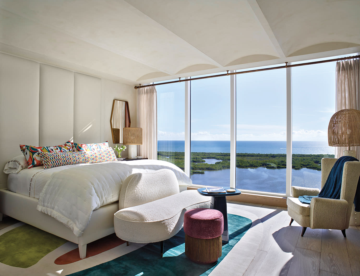 Bright, airy bedroom with large windows offering a serene ocean view. Features a plush bed, colorful cushions, and modern furniture on a geometric rug.