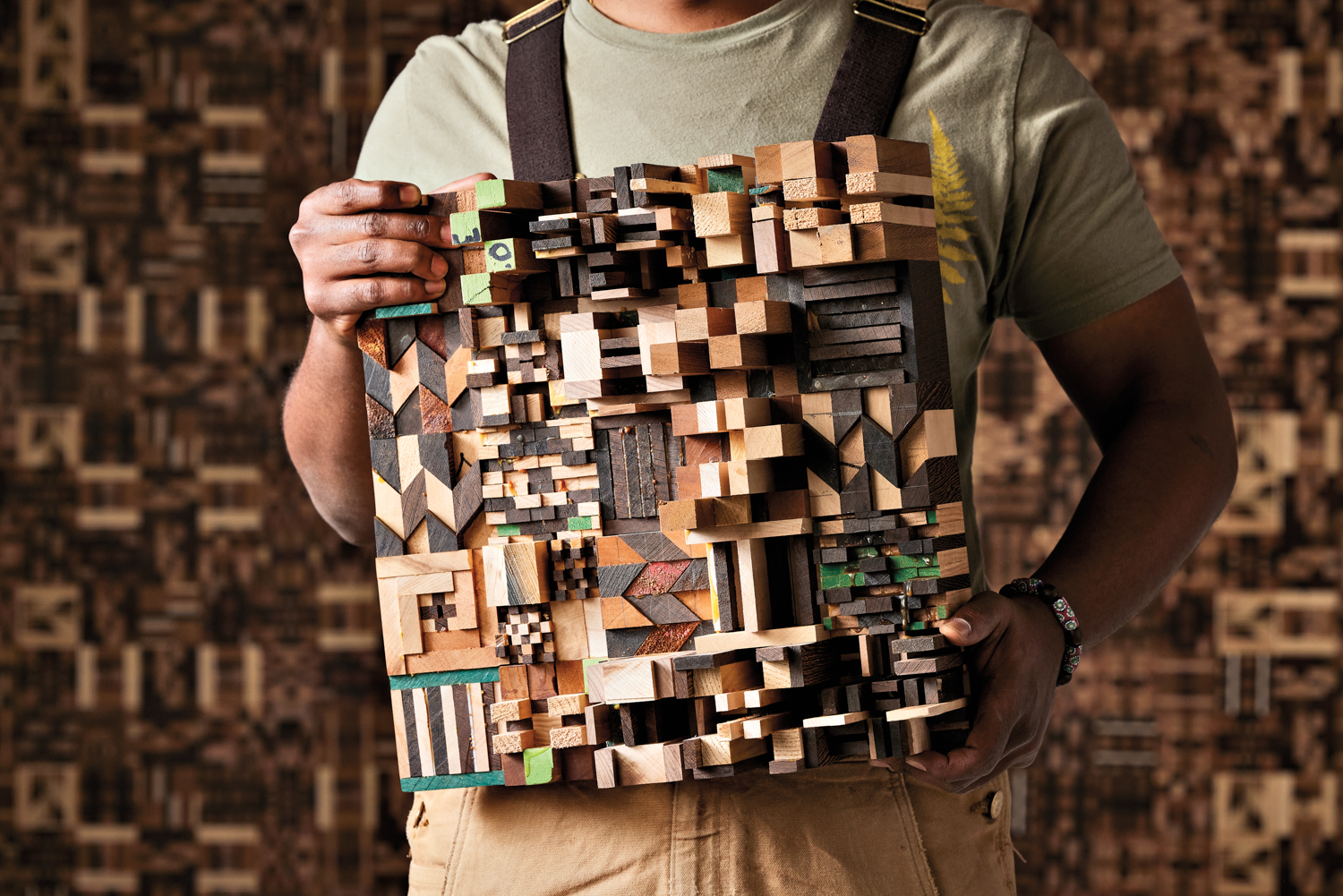 Ato Ribeiro stands in front of a patterned wood wall holding one of his wooden sculptures