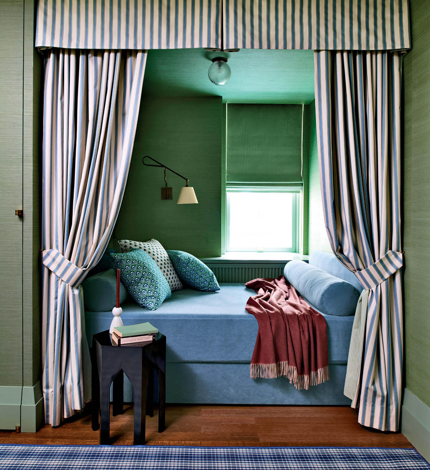 Cozy nook with a light blue daybed surrounded by striped curtains. Green walls, patterned cushions, and a pink throw create a soothing, intimate space.