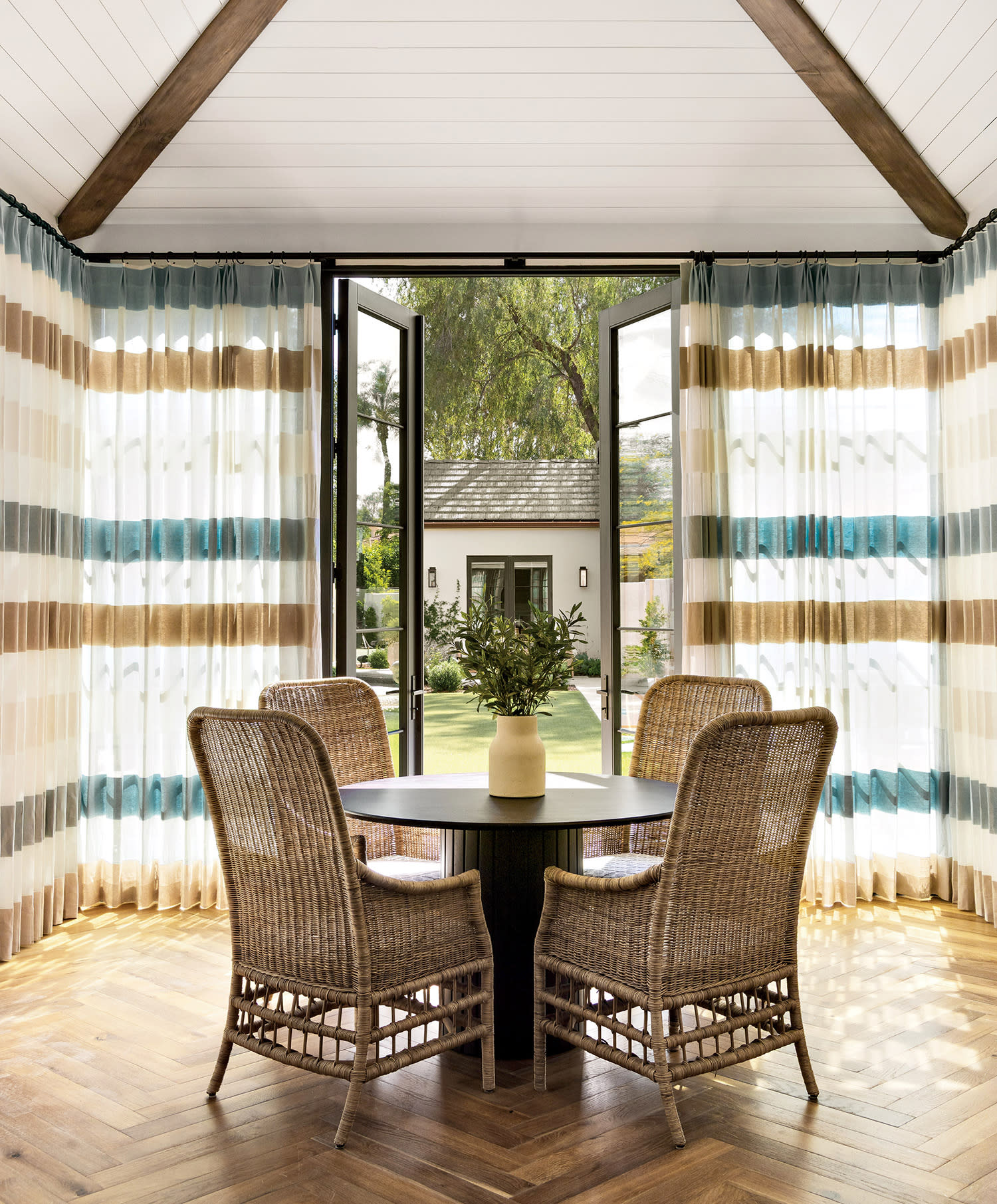 Four wicker chairs surround a round table with a vase, set in a room with herringbone wood floors, large windows, and striped curtains, overlooking a garden.
