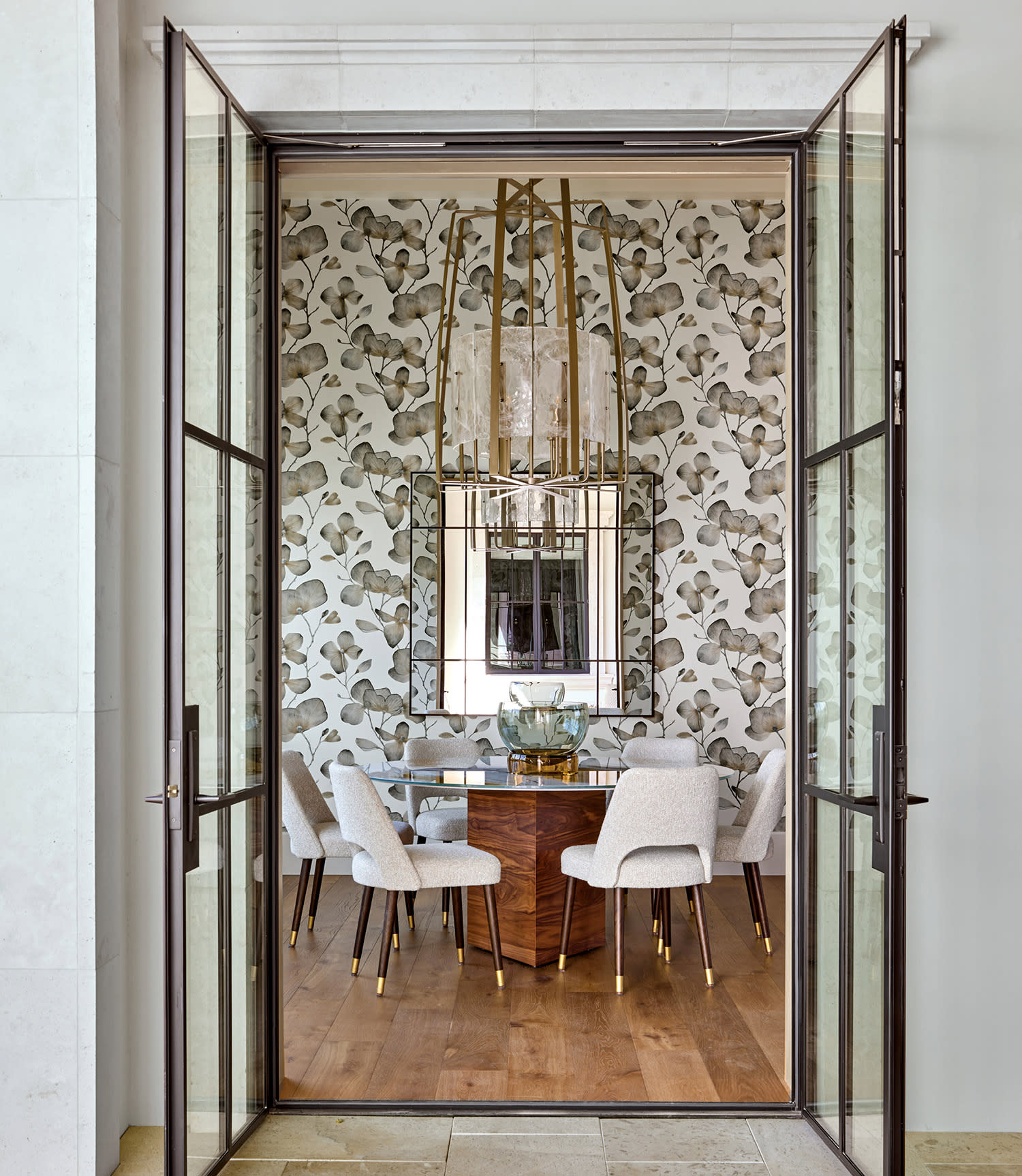 Elegant dining room with glass doors, floral wallpaper, and a wooden table surrounded by six white chairs. A chandelier above adds a touch of luxury.