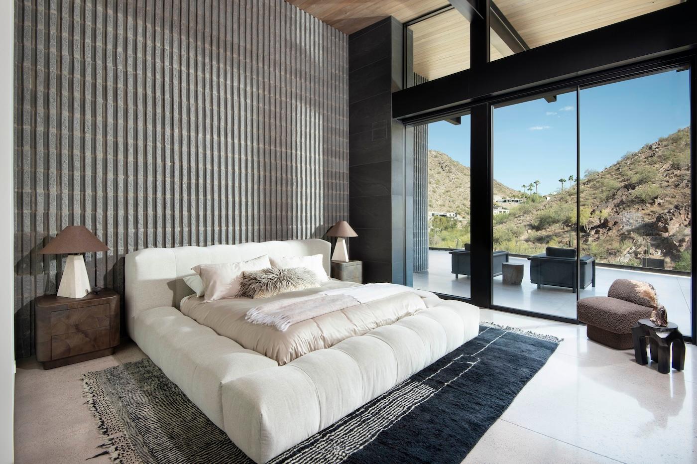 Modern bedroom with large bed, textured wall, and floor-to-ceiling windows overlooking a mountainous landscape. Neutral tones and minimal decor create a serene atmosphere.