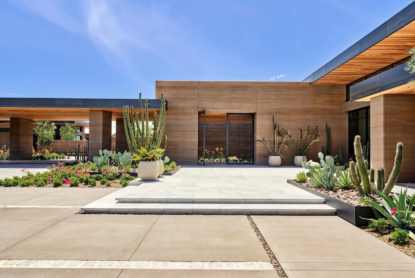 Modern, minimalist house with brown walls, large windows, and a flat roof. Cacti and succulents in planters decorate the garden under a clear blue sky.
