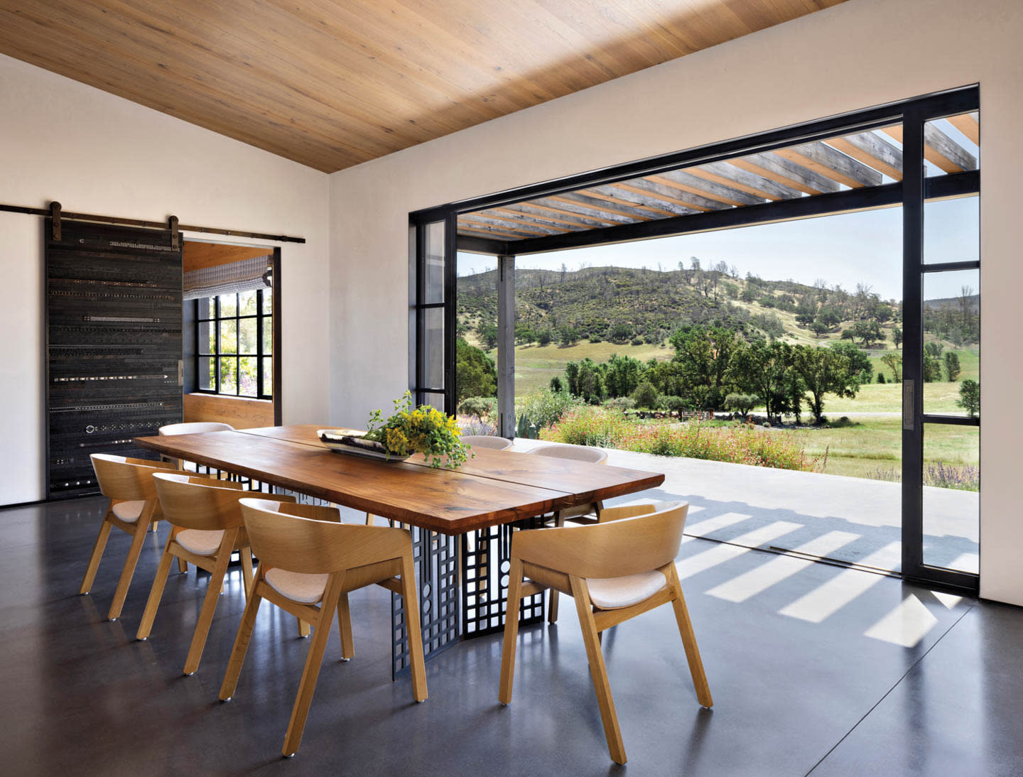 Modern dining room with a wooden table and chairs, large open sliding doors reveal a scenic view of rolling hills and greenery, creating a tranquil atmosphere.