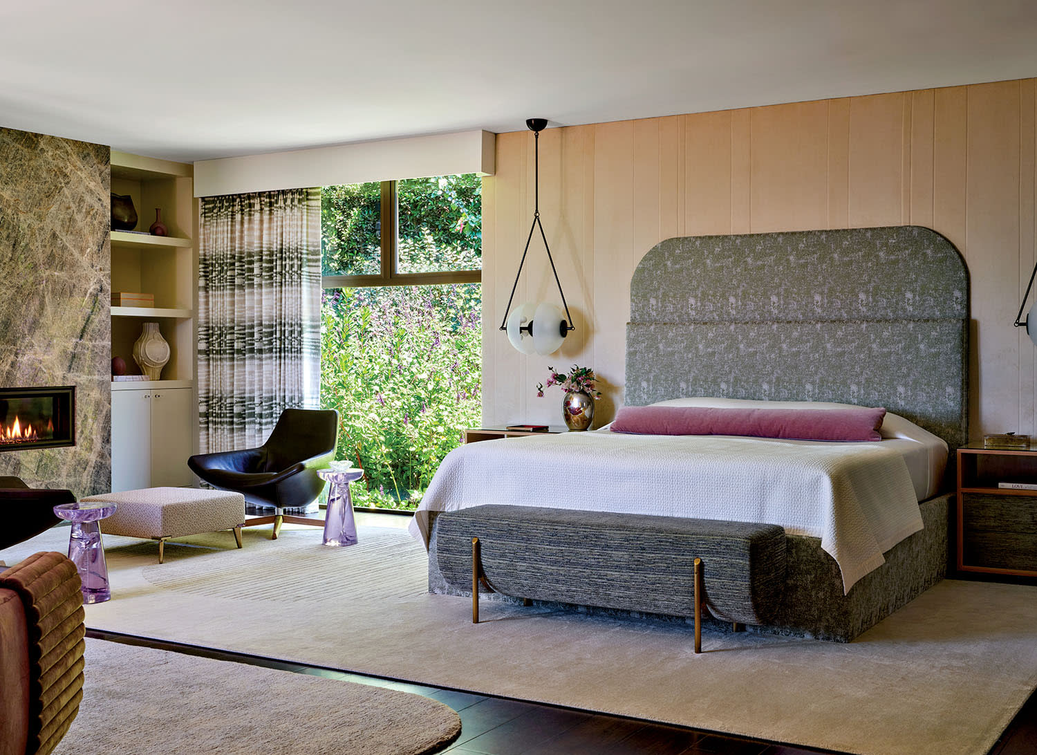 Modern bedroom with a large bed, gray headboard, and pink pillow. Features a fireplace, glass pendant lamps, and chair near a window with garden view.