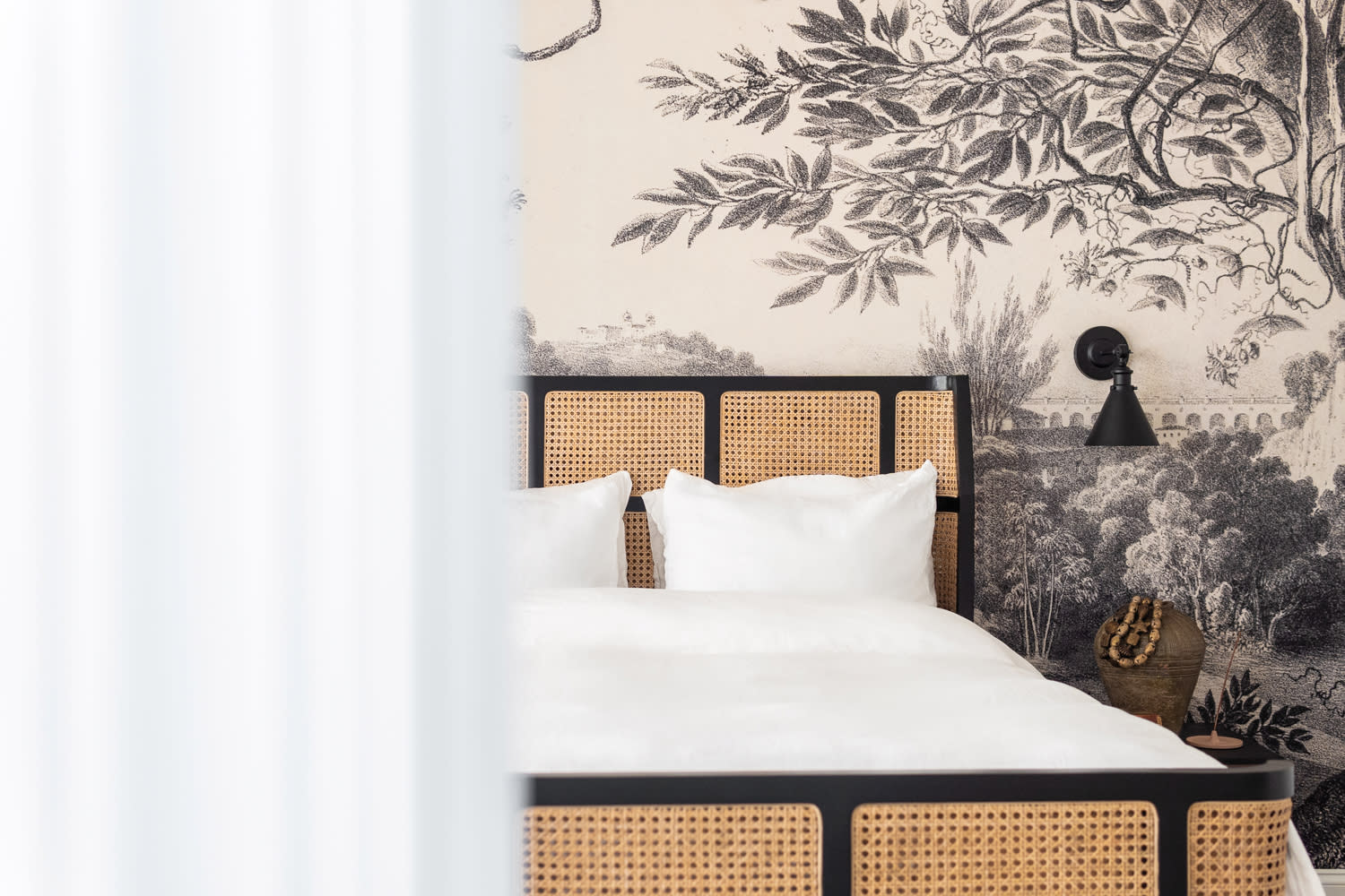 Black-and-white wallcovering of a landscape backs a wicker bed dressed in white linens.