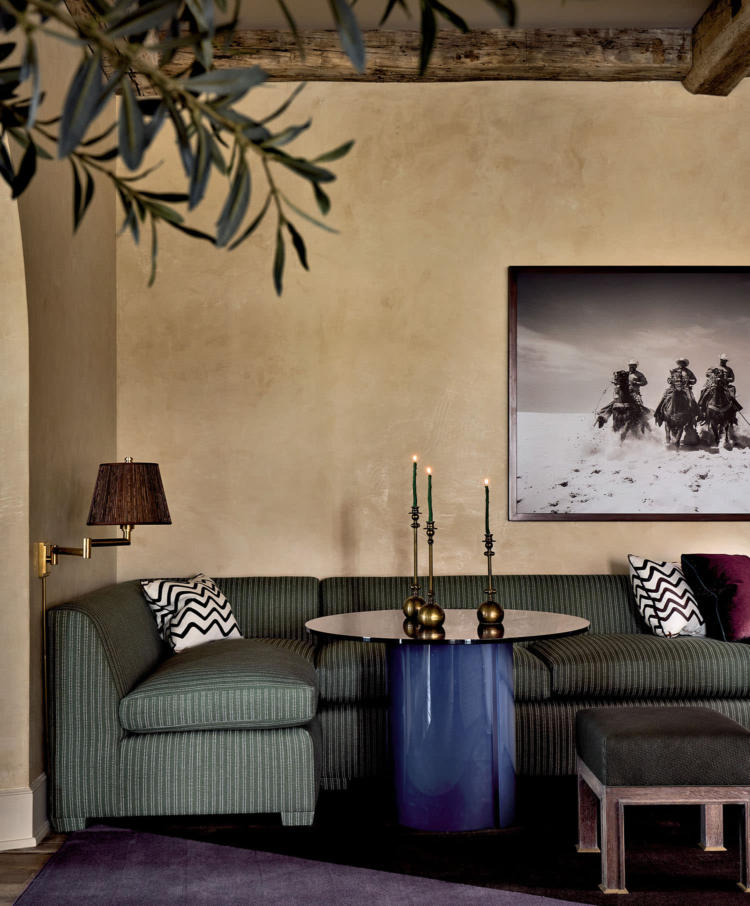 Stylish living room with green-striped sofa, blue round table, and dark candles. A black-and-white cowboy photo hangs on the beige wall. Cozy ambiance.