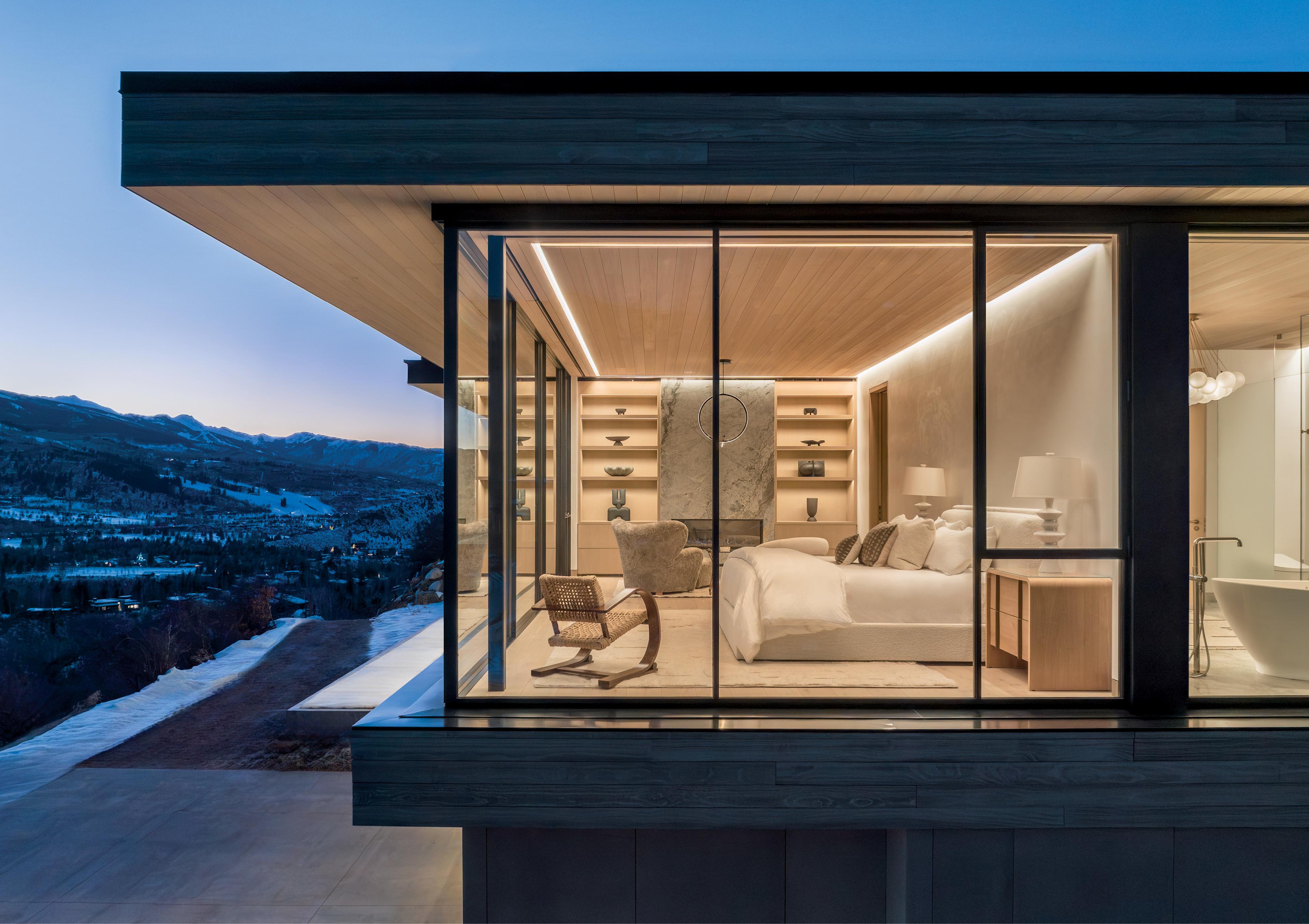 Modern bedroom with floor-to-ceiling windows showcasing a mountain view at dusk. Warm lighting and minimalist decor create a cozy ambiance.