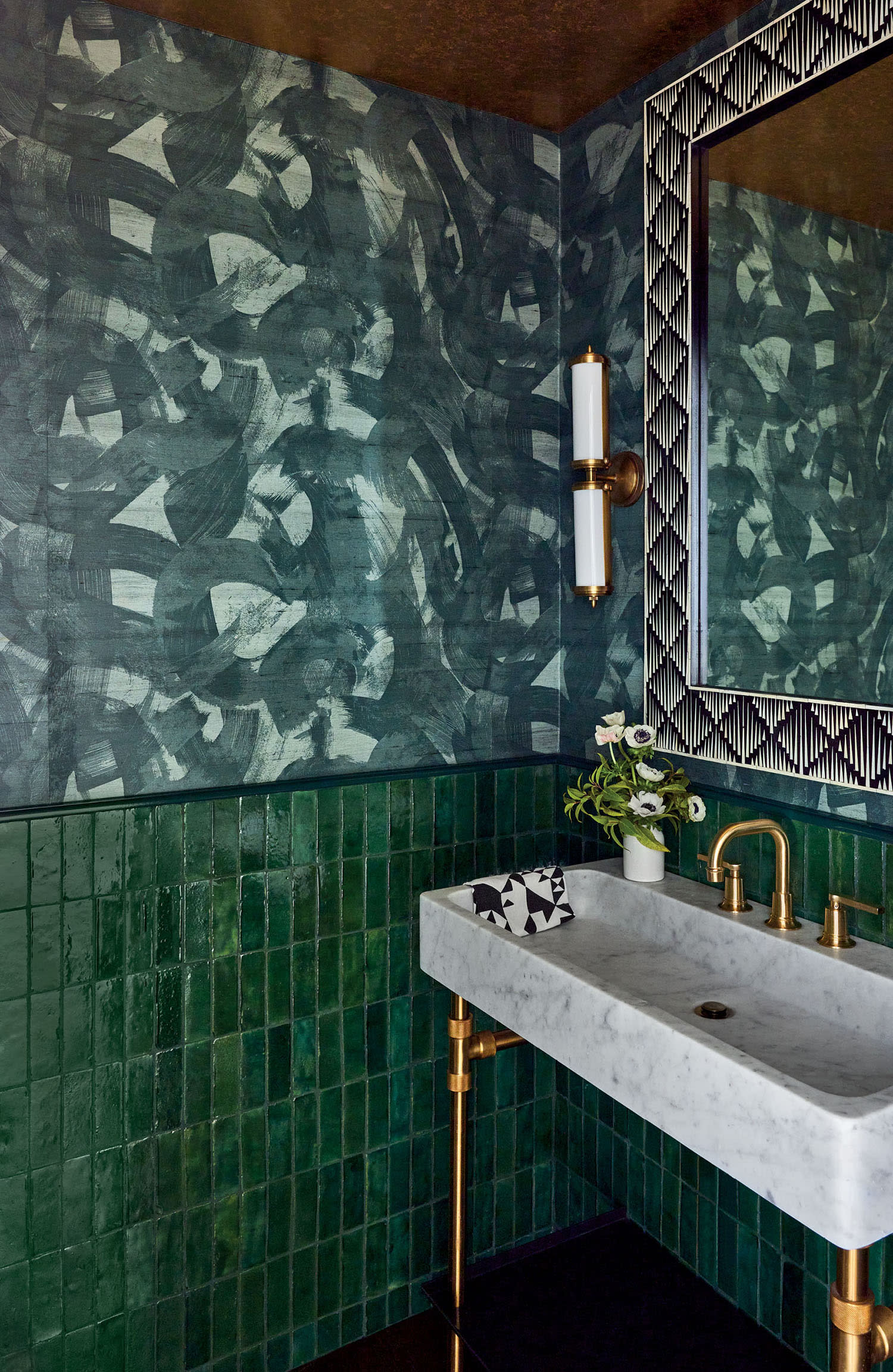 A bathroom with green tile, patterned wallpaper and a standing sink vanity under a mirror.