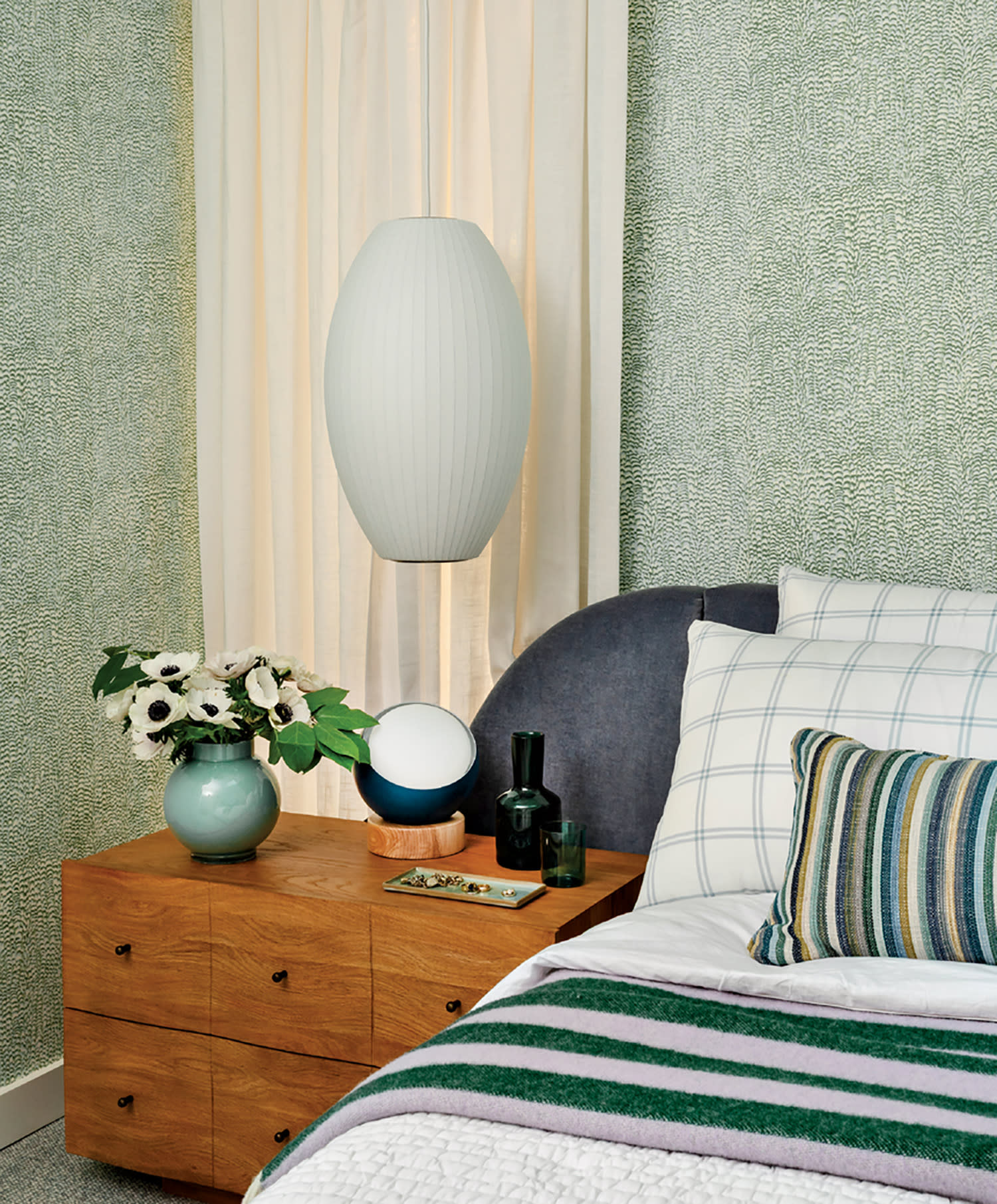 Cozy bedroom with green patterned wallpaper and soft lighting. A wooden nightstand holds a vase with flowers, decorative objects, and a bed with striped pillows.