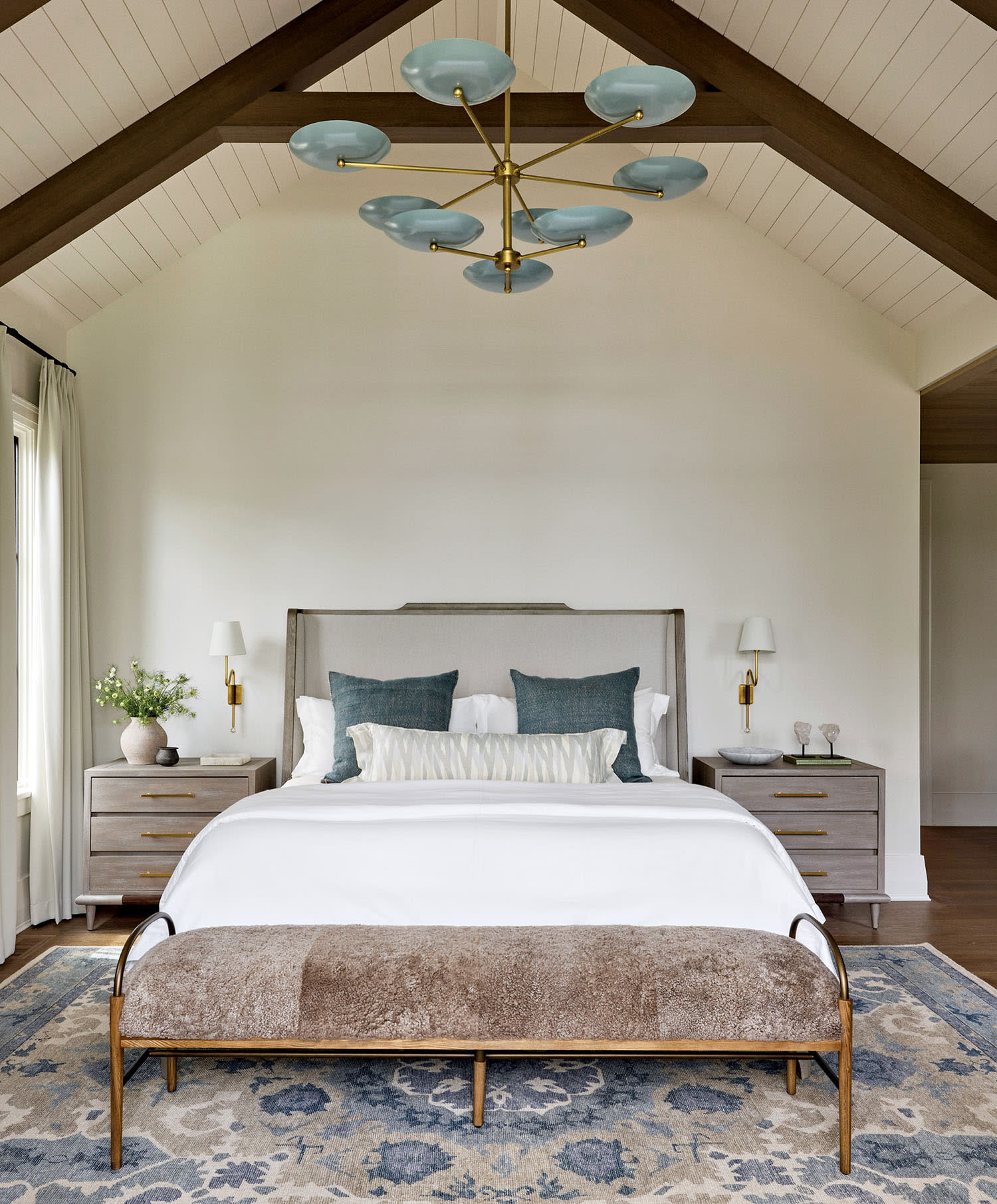 Spacious bedroom with vaulted ceiling and modern chandelier. Features a bed with blue cushions, white bedding, and a plush bench, creating a cozy ambiance.