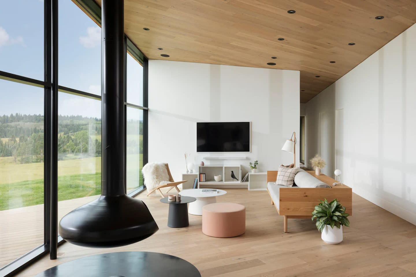 Modern living room with a large window, wooden ceiling, sleek black hanging fireplace, minimalist furniture, TV, and soft tones. Bright and serene.