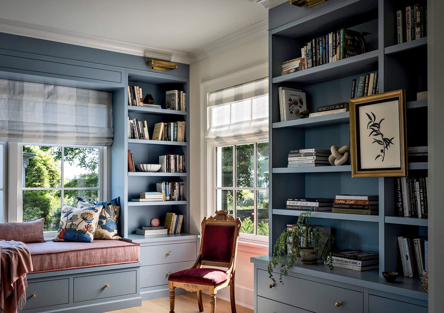 Cozy reading nook with blue built-in bookshelves, a window seat, and a red velvet chair. Soft light filters through the windows, adding warmth.