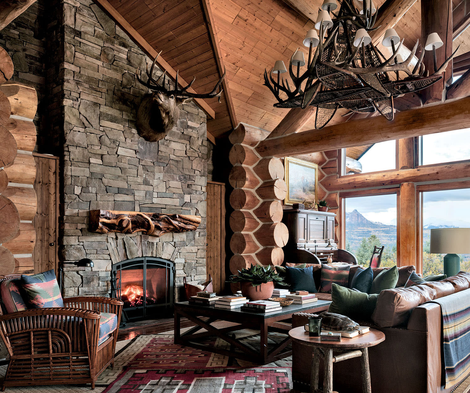 A room with wood walls, a stone fireplace and large windows, decorated with a couch, accent table and armchair.