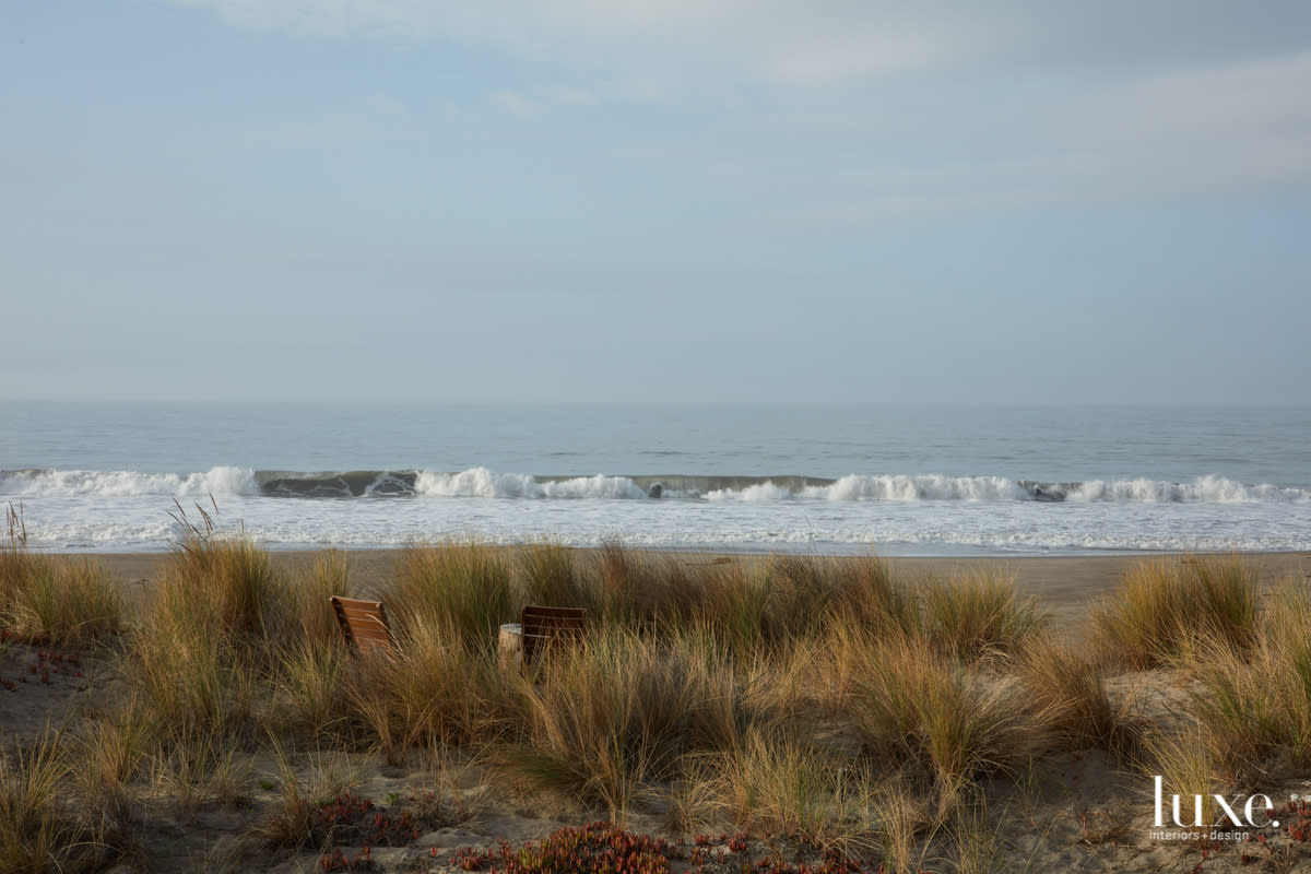 Two chairs seemingly hidden in sand dune grass face the periwinkle shoreline.