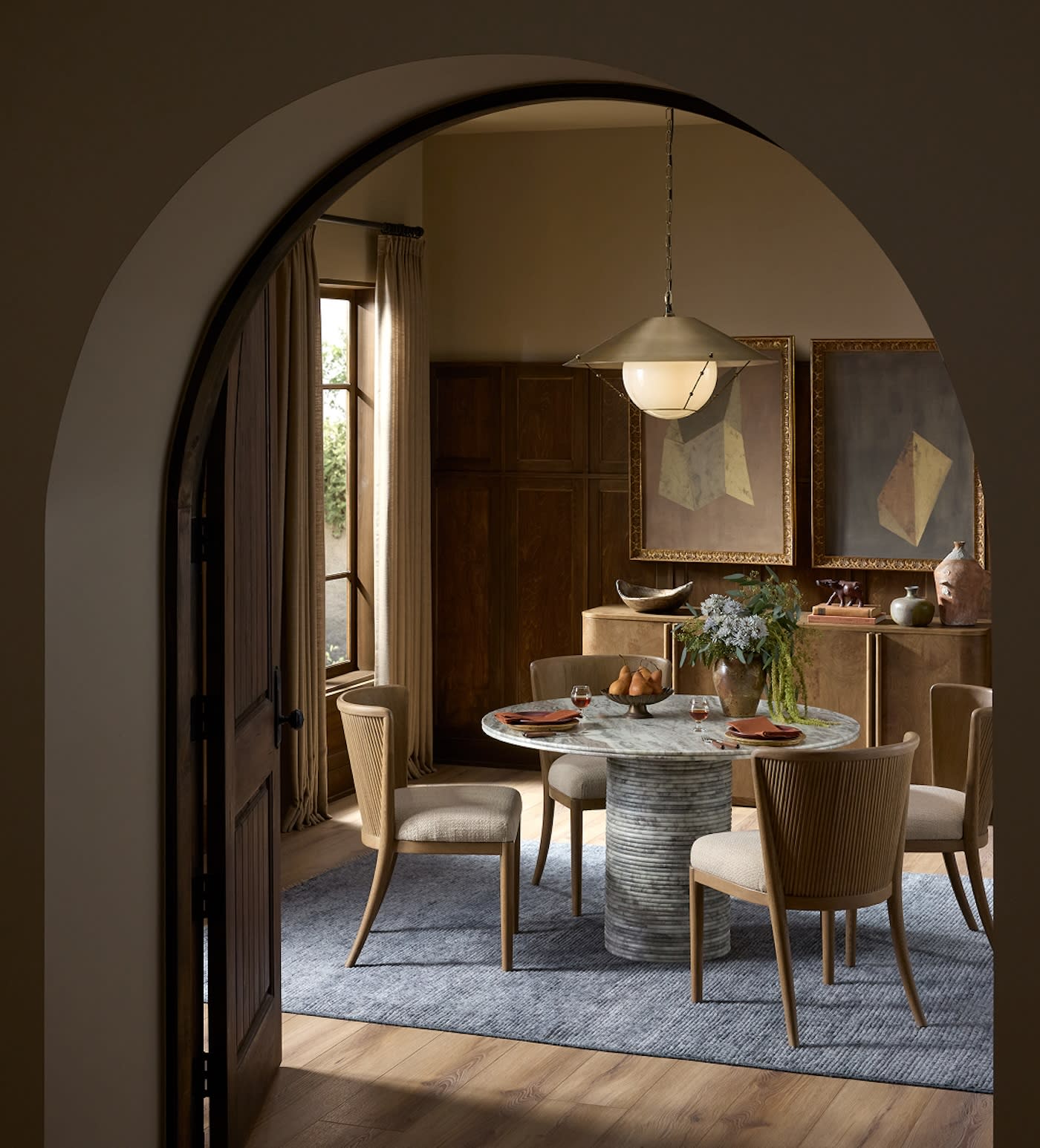A cozy dining room with a round marble table, four wooden chairs, and a textured blue rug. Soft lighting, art on walls, and a serene, inviting ambiance.