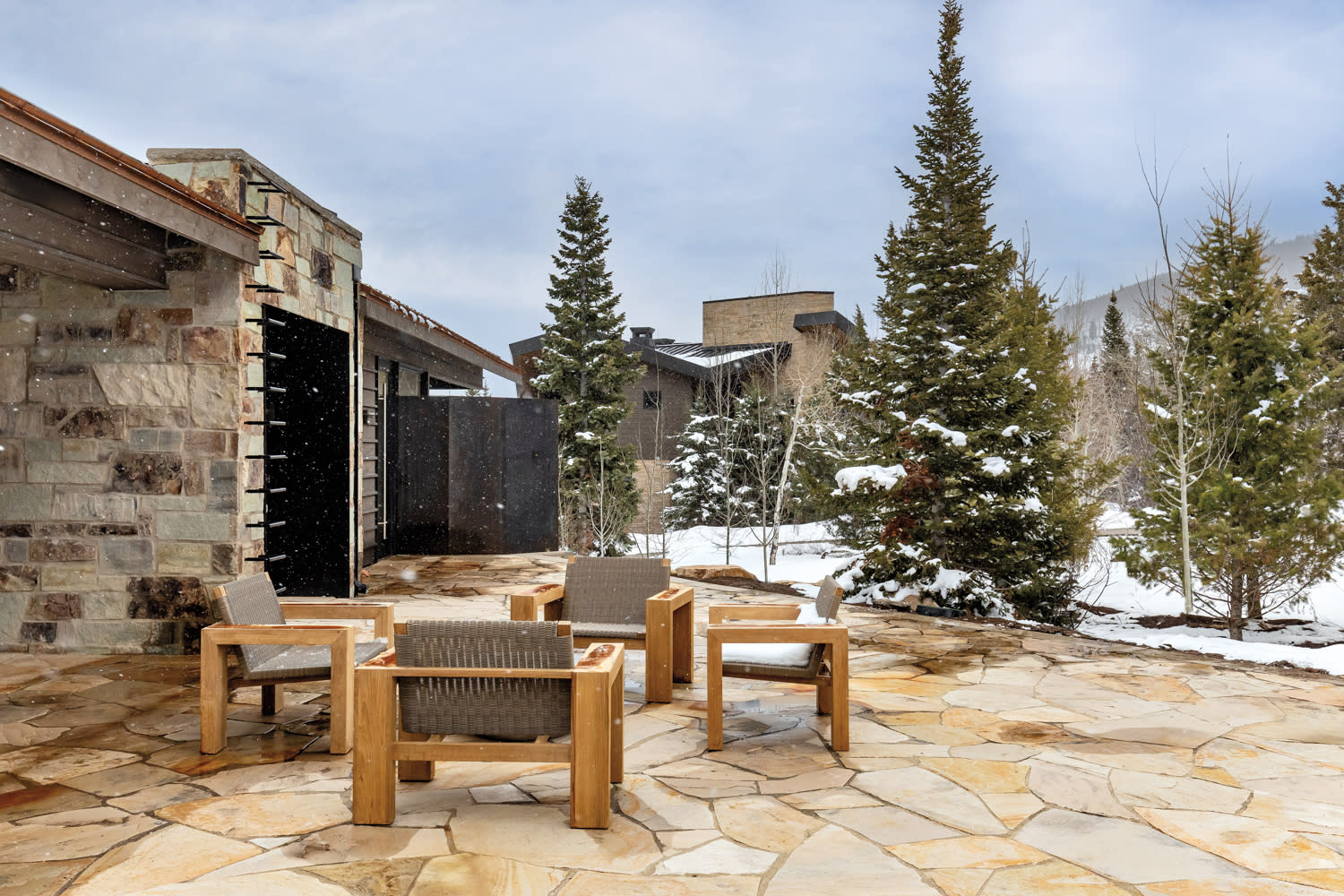 Snow-dusted patio with wooden chairs around a stone table, set against evergreen trees and a stone building. The scene feels peaceful and wintry.