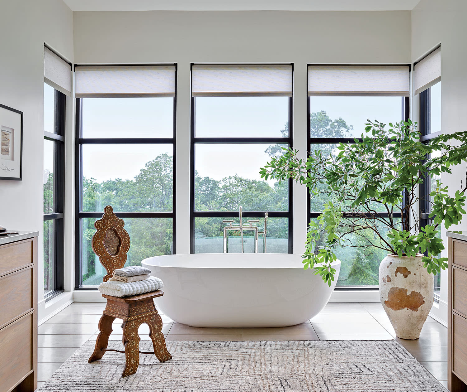 A freestanding tub in a bathroom with an accent chair, vase with greenery, and large windows.