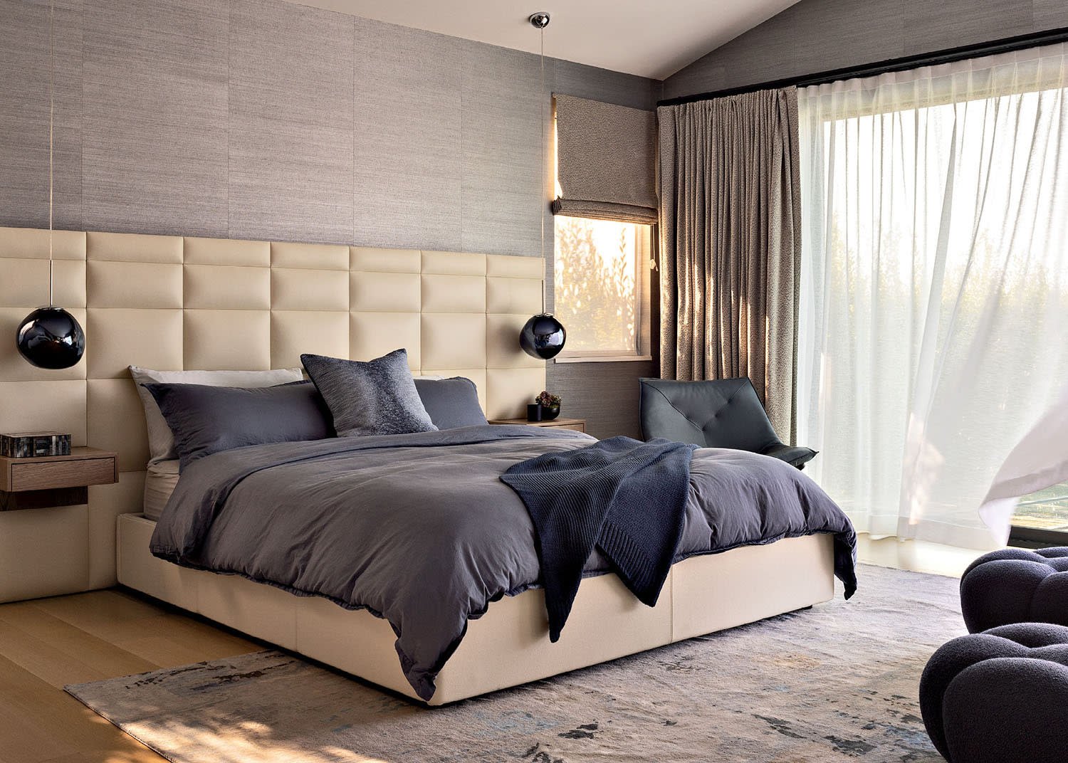 A white leather headboard sits behind a bed with dark linens alongside a large window.