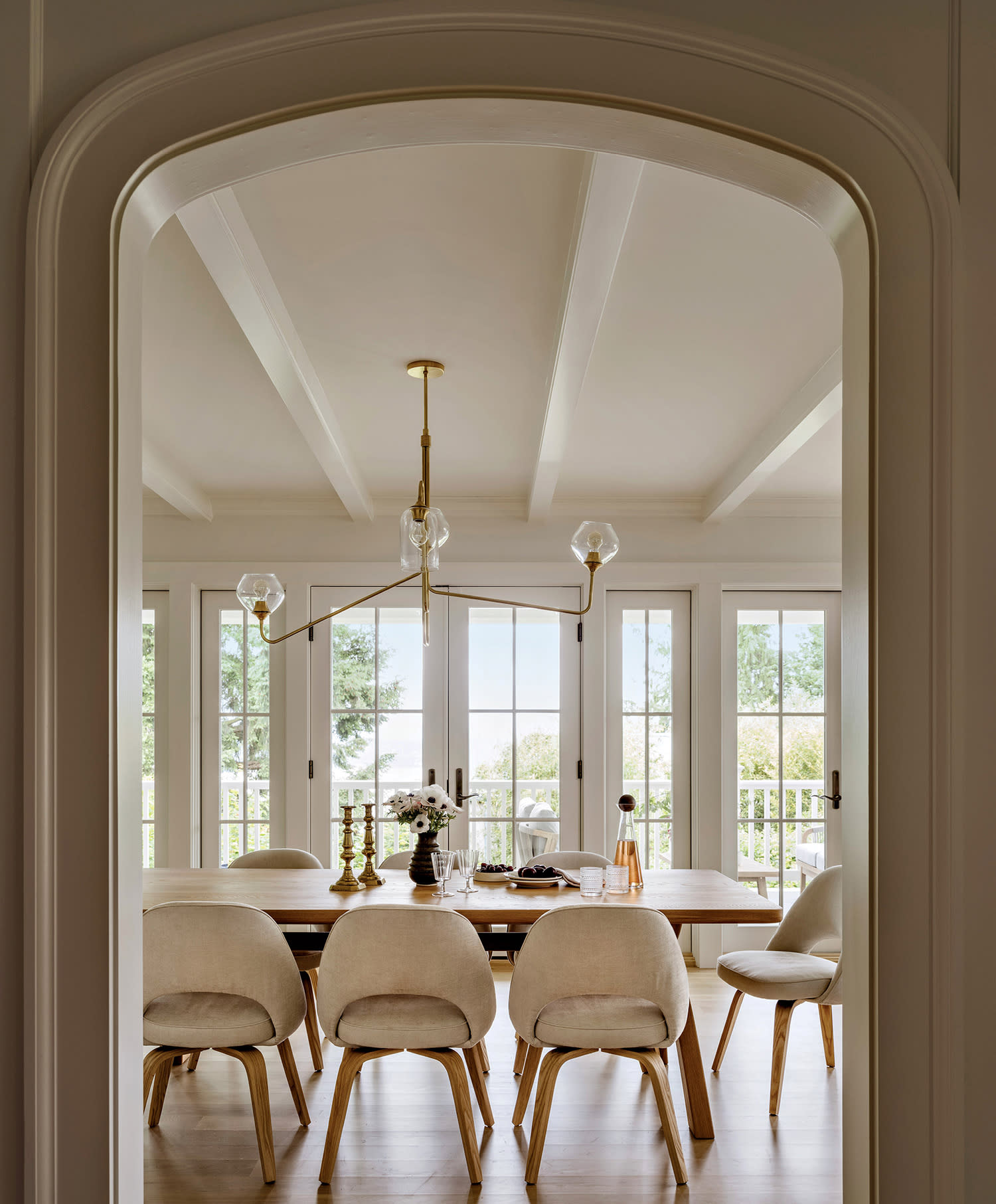 Elegant dining room with a wooden table and eight beige chairs, set beneath a modern chandelier. Large windows offer a view of greenery outside.