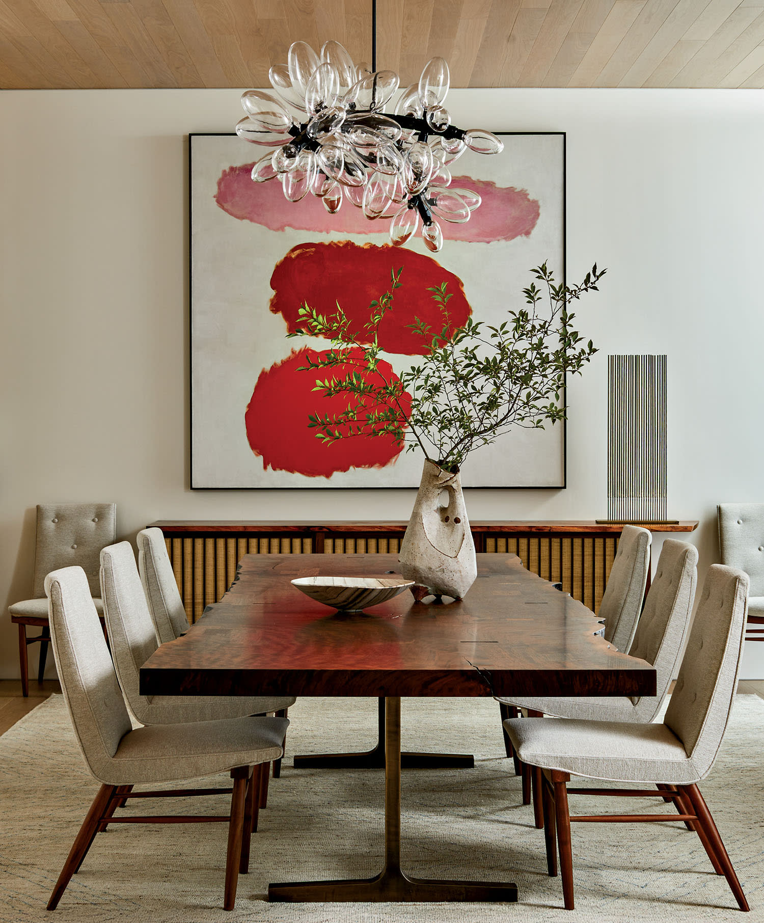 Sophisticated dining room with a large wooden table, six beige chairs, abstract red painting, ornate chandelier, and leafy plant centerpiece.