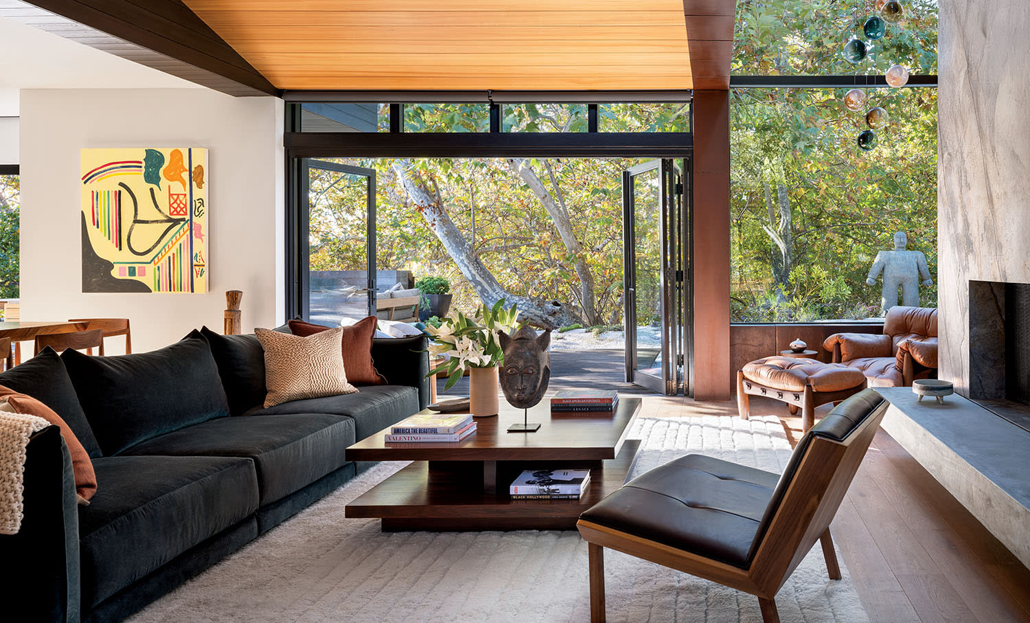 Modern living room with large windows and natural light. Features a dark sofa, wooden coffee table with decor, abstract artwork, and view of lush trees outside.