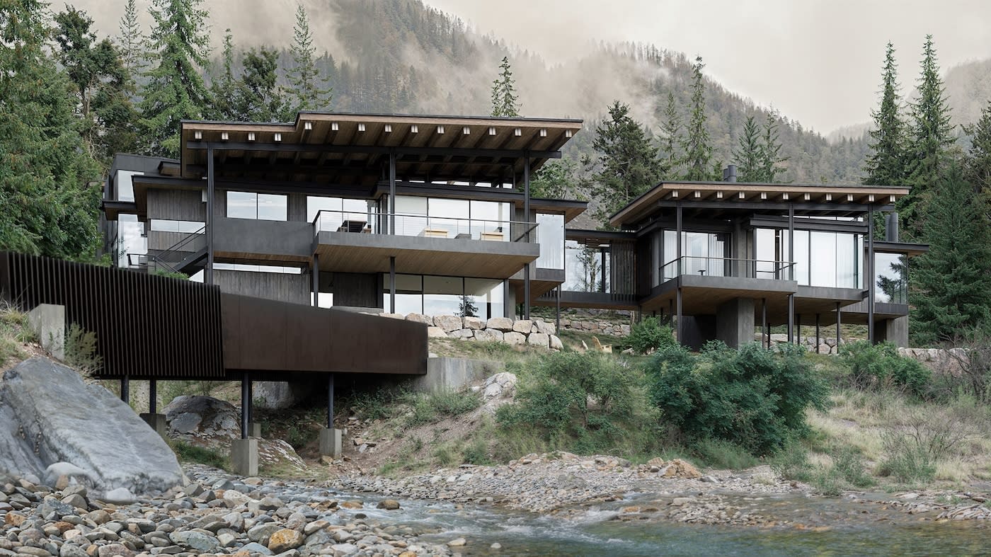 Modern homes with large glass windows are nestled in a lush mountain landscape, surrounded by trees and a flowing stream, creating a serene atmosphere.