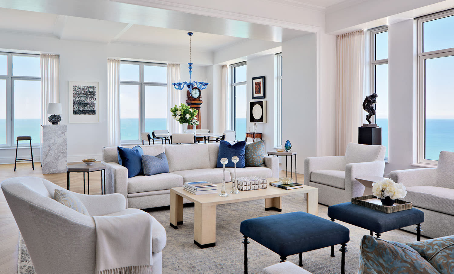 White couches and armchairs set around a light-colored wood coffee table with blue ottomans nearby.