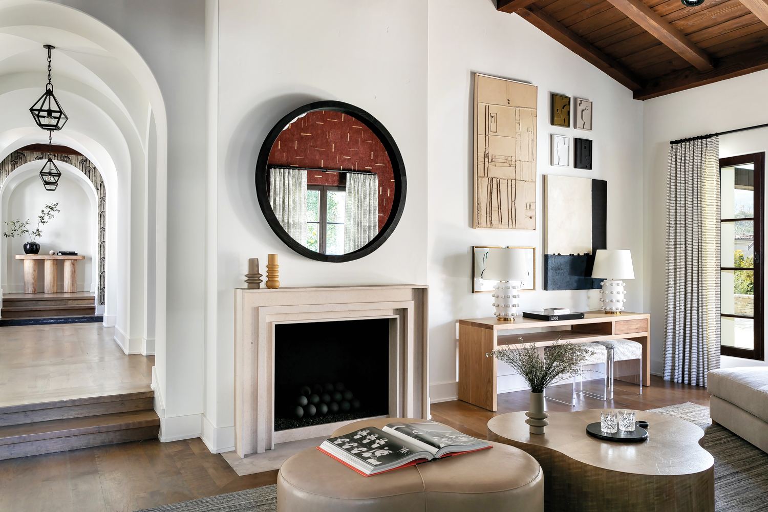 Living room with round, black-framed mirror above fireplace, natural color palette throughout and assortment of abstract paintings