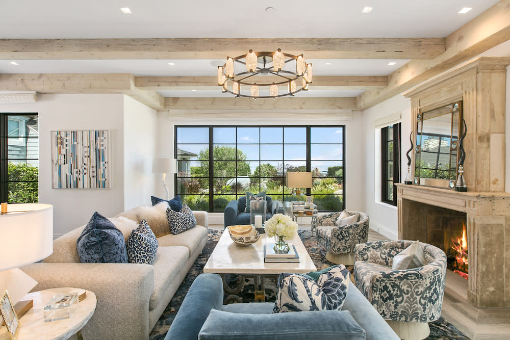 A modern living room with beige sofas, patterned blue chairs, a marble coffee table, a fireplace, a chandelier, and large windows overlooking greenery decorates the space.