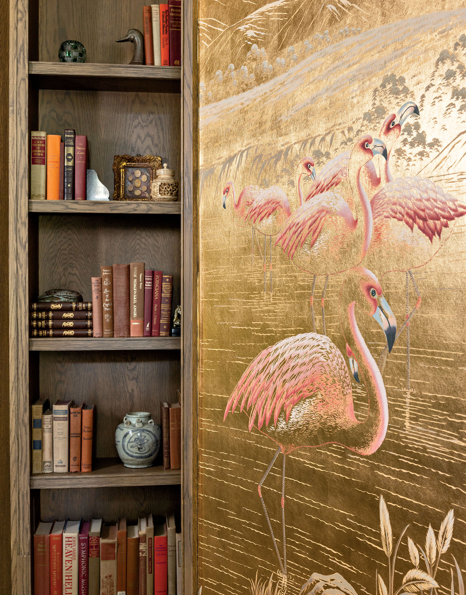 Bookshelf filled with colorful books and decorative items next to an ornate gold panel featuring painted flamingos. The scene exudes elegance and creativity.