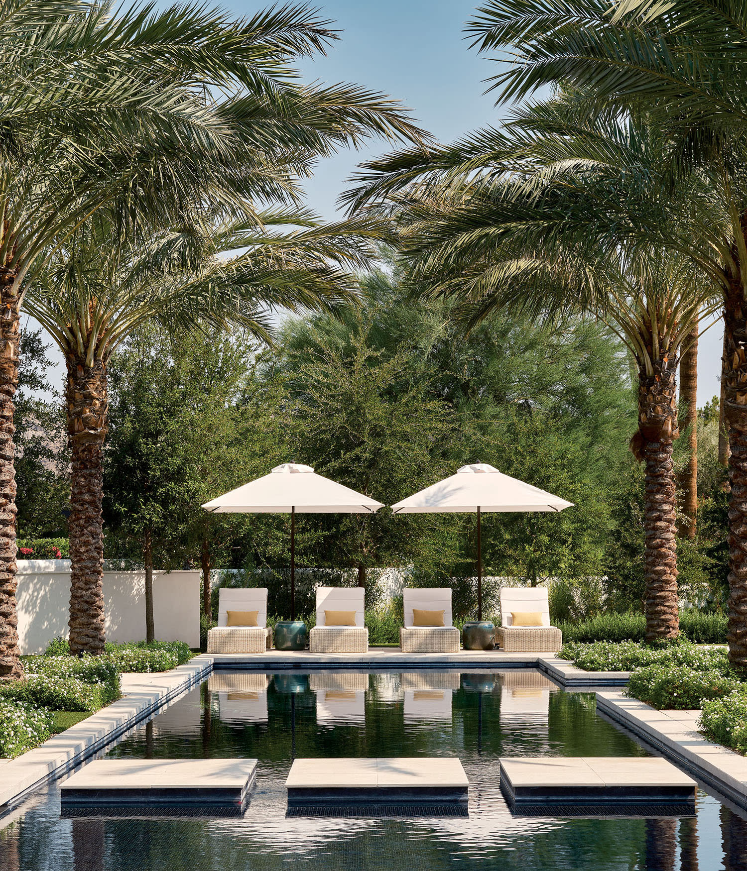 Luxurious pool with palm trees, three lounge chairs under umbrellas, and manicured greenery. The scene conveys a tranquil, resort-like atmosphere.