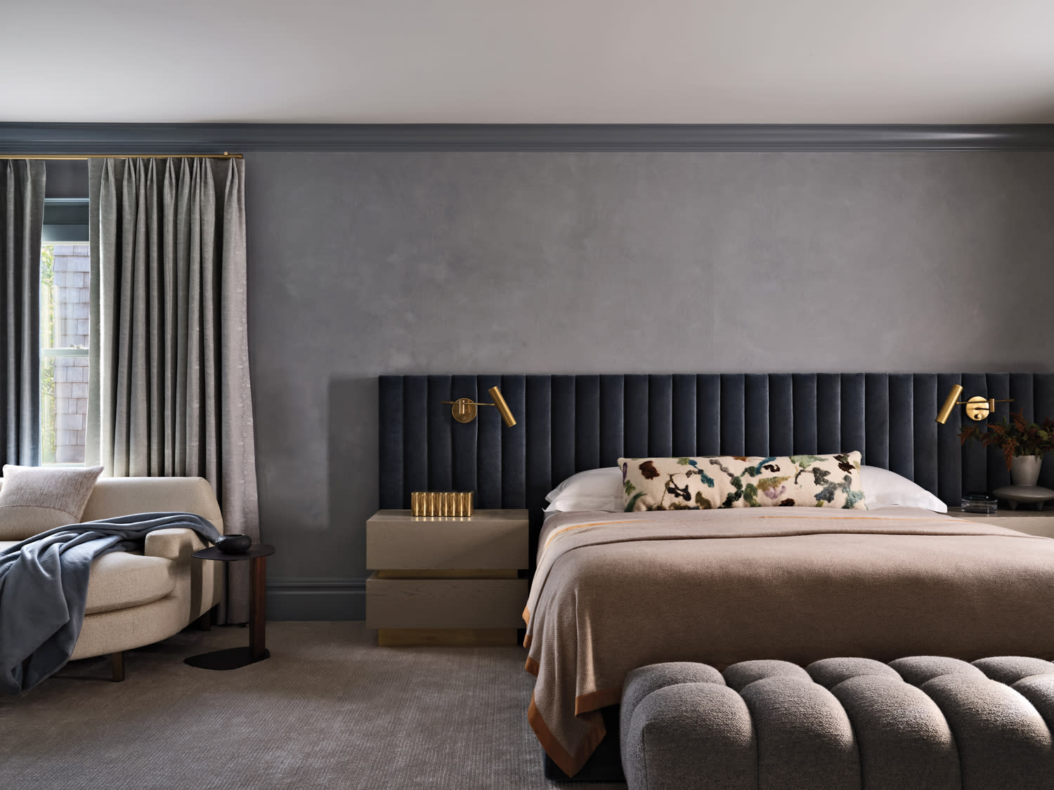 Modern bedroom with gray textured walls, a plush navy headboard, and beige bedding. A cozy armchair and draped curtains frame a window, exuding comfort.