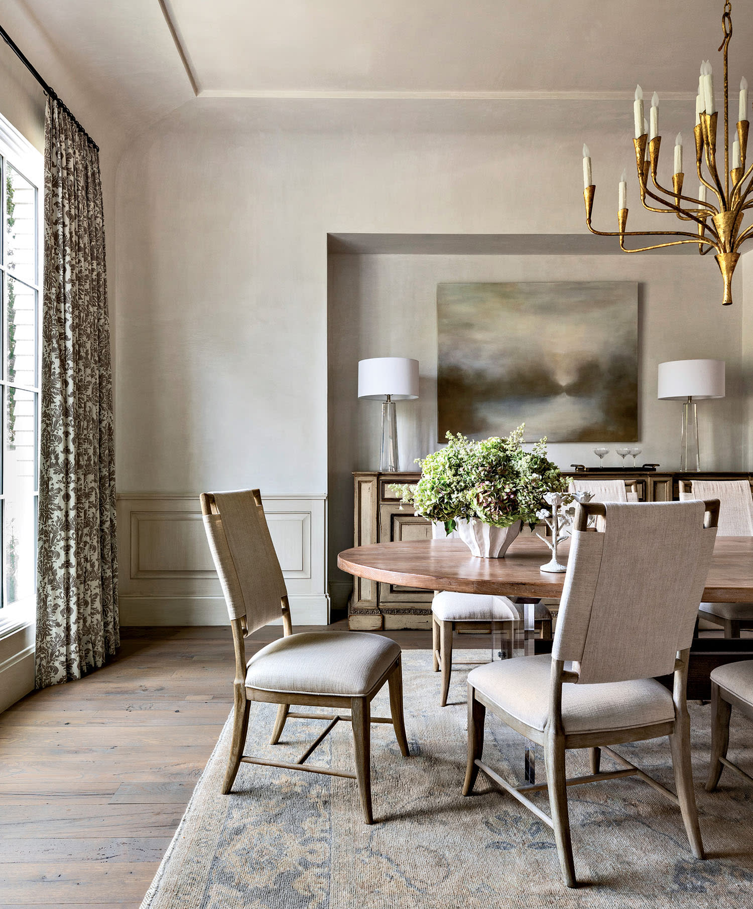Elegant dining room with a round wooden table, four upholstered chairs, and a centerpiece of greenery.