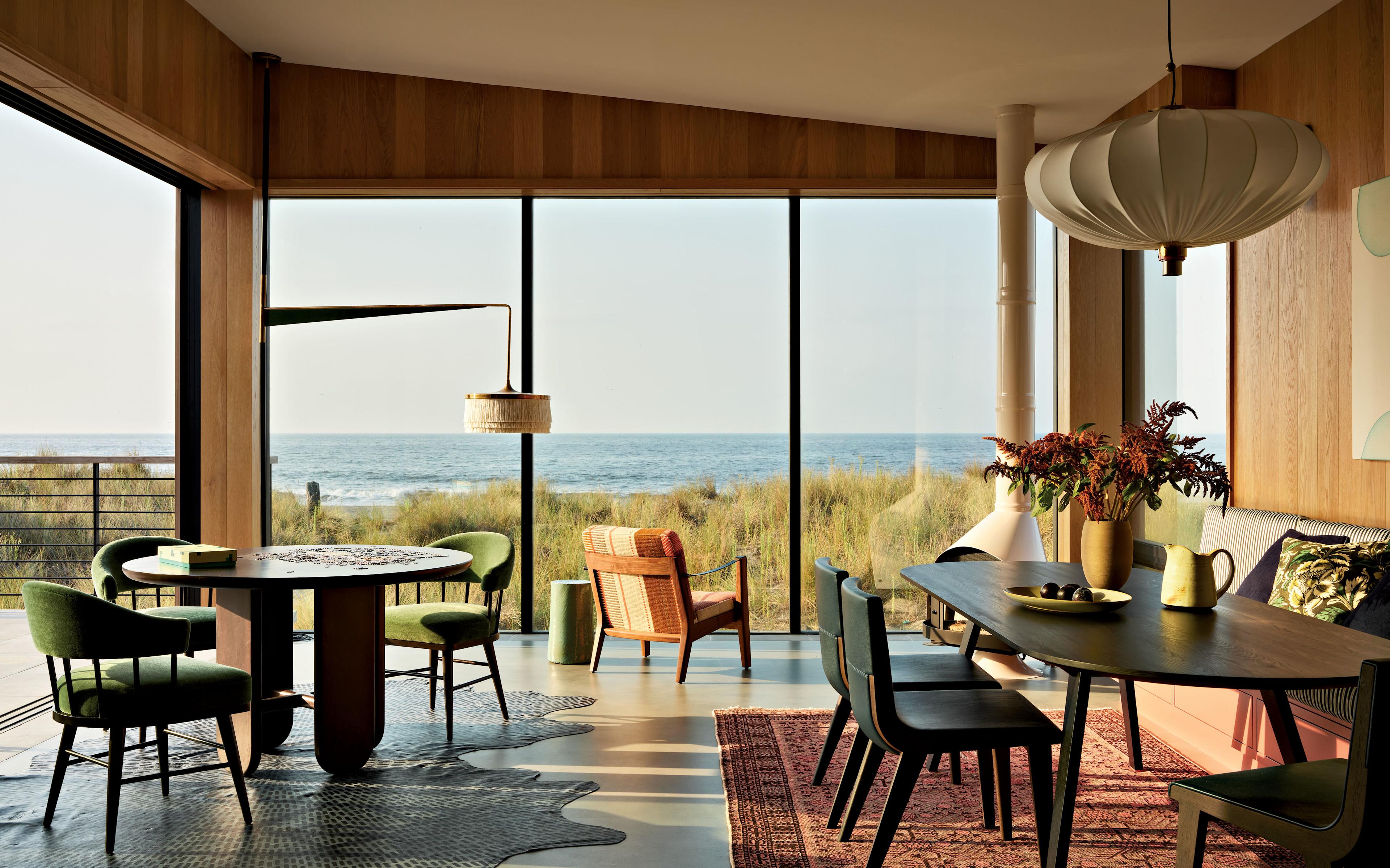 Stylish coastal living room with large windows overlooking the ocean. Features a round table, green chairs, wicker chair, and pendant lighting. Warm, inviting ambiance.