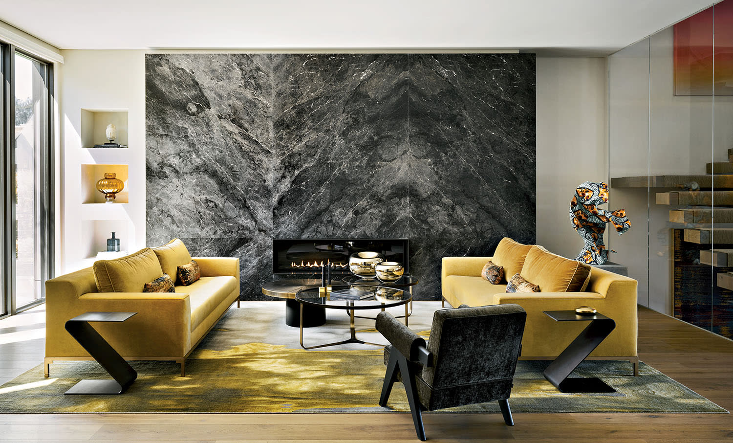Modern living room with mustard sofas, a sleek black armchair, and a geometric coffee table. A striking marble fireplace wall adds elegance. Bright and inviting.