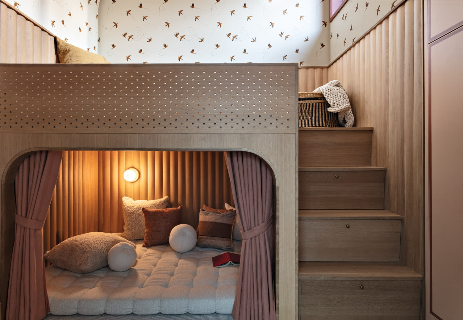 Cozy wooden nook bed with soft lighting, pillows, and a book underneath a staircase. Bird-patterned wallpaper adds a whimsical touch.