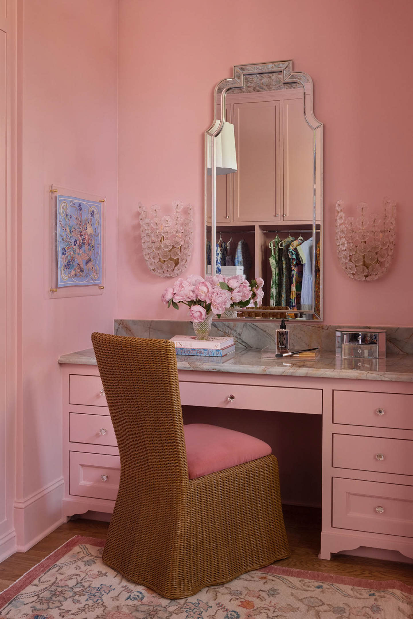 Pink vanity area with a wicker chair and marble countertop. A mirror reflects a closet; floral decor and books add elegance. Walls are soft pink.
