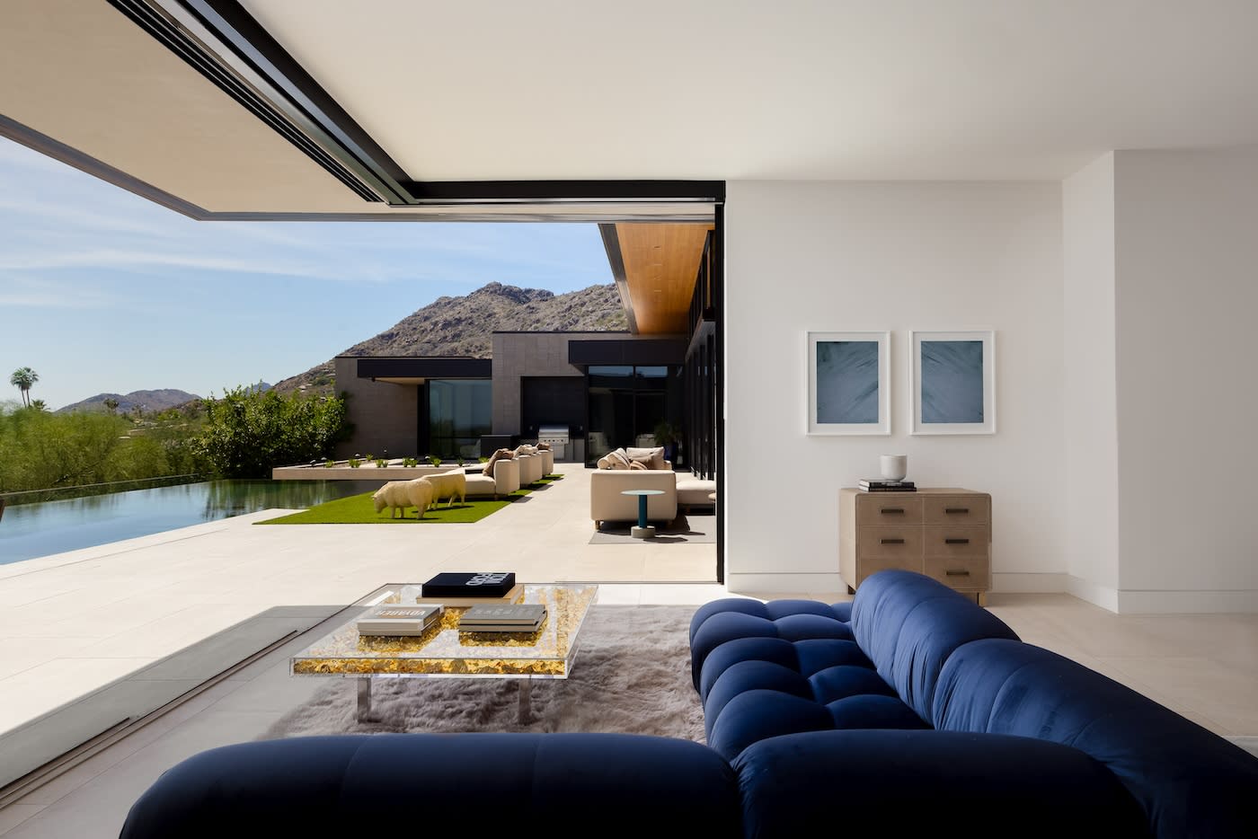 Modern living room with a plush blue sofa facing a glass coffee table. Open sliding doors reveal a sunny patio, garden, pool, and scenic mountain view.