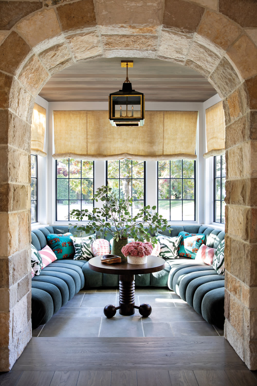 Breakfast nook seen through a stone arch, with a blue-velvet ribbed banquette and a round table with a geometric base.