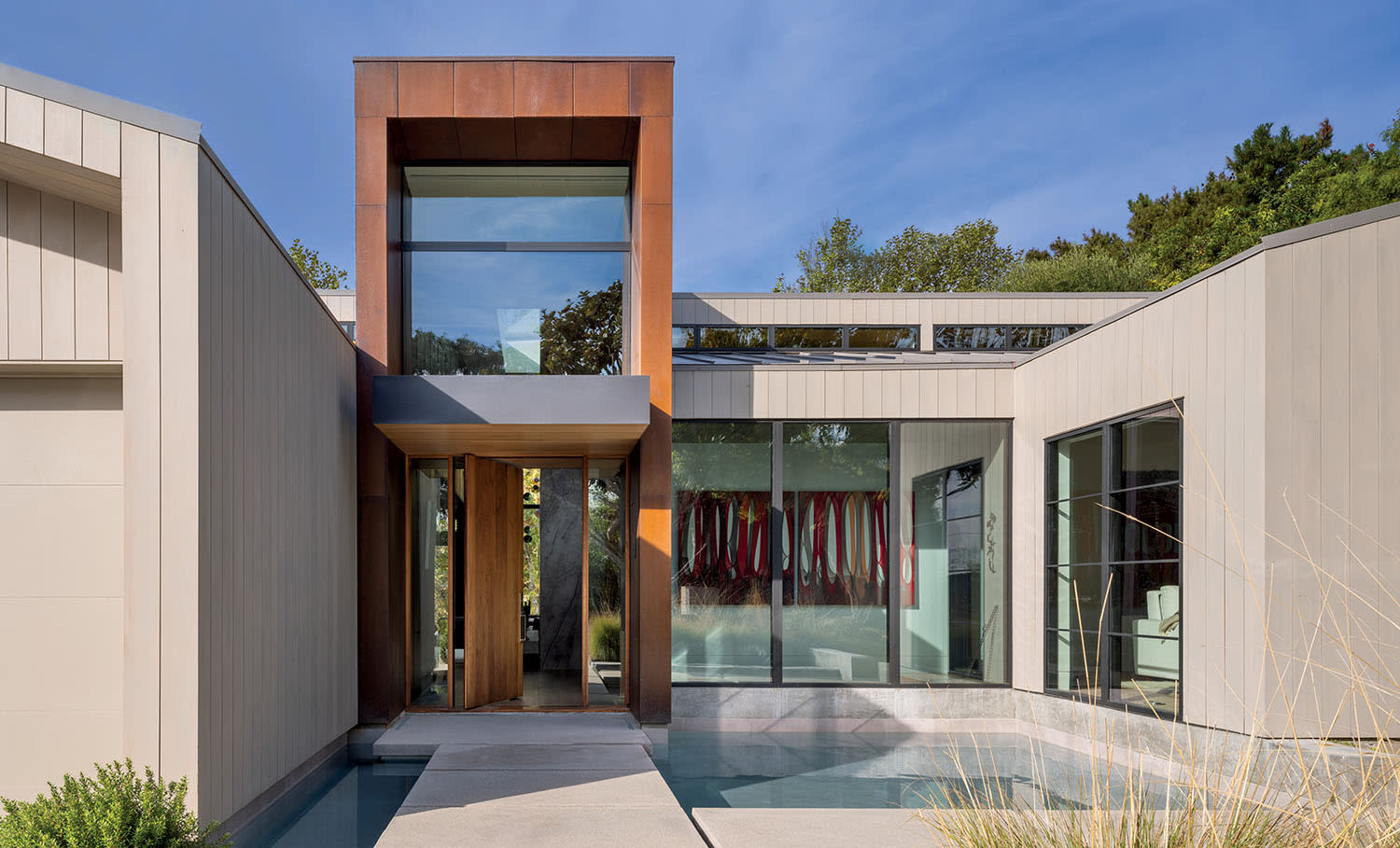 Modern house exterior with large windows, rust-colored metal accents, and a serene reflecting pool. Surrounded by greenery under a clear blue sky.
