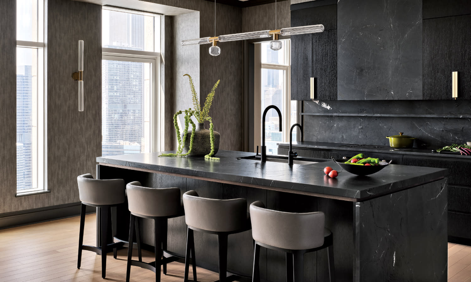 Stylish kitchen with sleek black marble countertops, modern pendant lights, gray chairs, and tall windows. A potted plant and fresh vegetables add color.