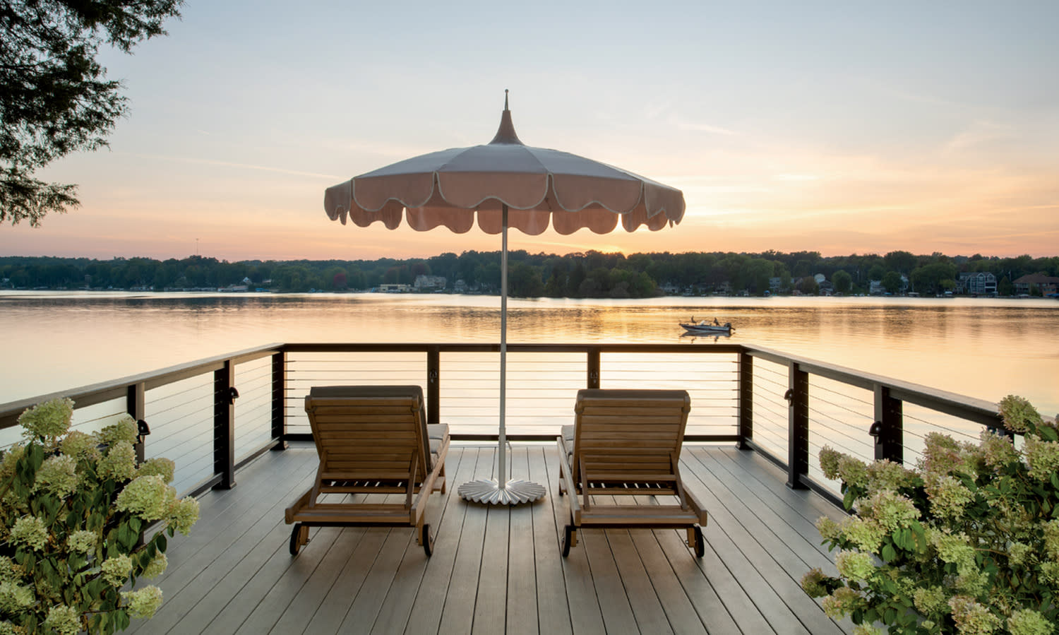 Two wooden lounge chairs under a large umbrella face a serene lake at sunset. The deck is bordered by greenery, creating a peaceful, relaxing ambiance.