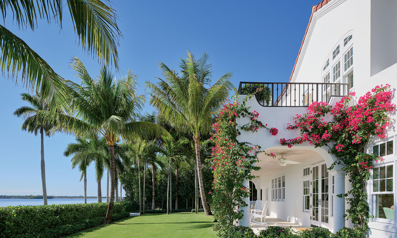 White house with lush greenery and vibrant pink climbing flowers, set beside a manicured lawn and tall palm trees under a bright blue sky.