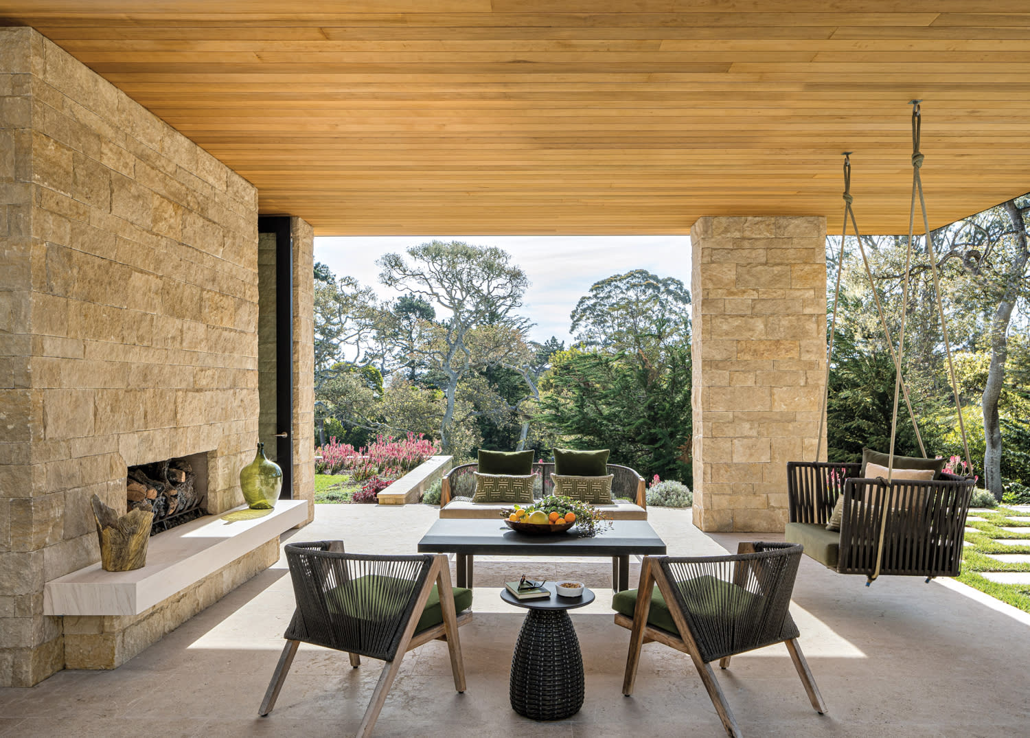 A shaded outdoor patio has seats around a fireplace, while the sofa and chairs invite people to relax