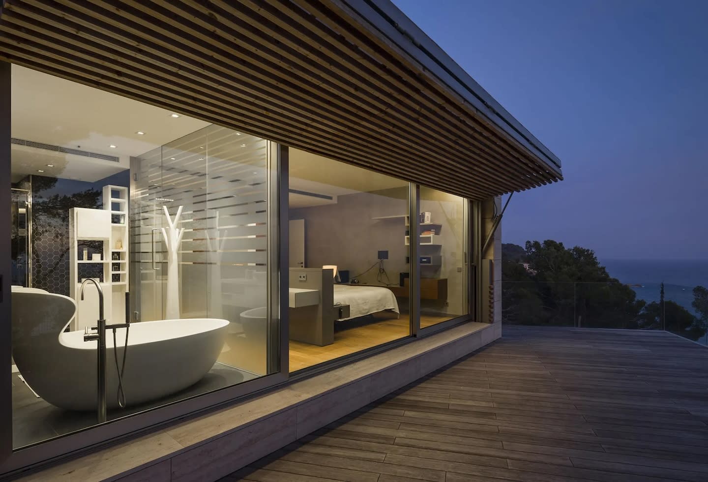 Modern glass-walled room at dusk with a free-standing bathtub and a bed. The space is calm, overlooking a serene ocean view, conveying relaxation.